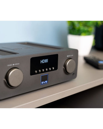 SVS Prime Wireless Pro Soundbase, aparatul pe o masă de lemn, cu o boxă pasivă în fundal și telecomanda alături
