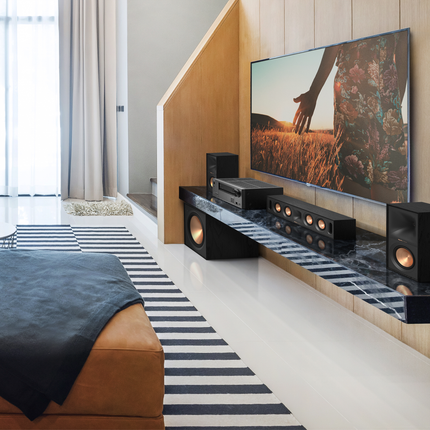 Boxa Klipsch R-50M fara grila pe o consola sub un televizor, langa un receiver AV si un canal central