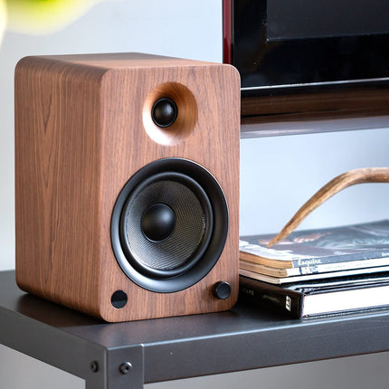 A Kanto Audio YU6 speaker in walnut on a black console, next to a stack of magazines